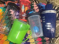 Photo showing six liquid containers: three colorful plastic mugs including a green handled mug and an orange mug with ice cream graphics, a clear measuring cup, a protein shaker with blue lid, and a blue martini shaker.