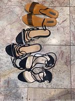 Three pairs of women’s shoes laid out on a light marble floor including two pairs of black heeled sandals and one tan sandal.