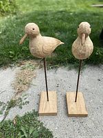 Two carved wooden bird sculptures with glass eyes mounted on steel rods and square wooden bases, standing outdoors on concrete with grass background.