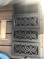 Five floor registers; one chrome finish metal with adjustable louvers and four black decorative metal with ornate cut-out grille patterns arranged vertically