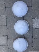 Three round white frosted glass flush mount light fixtures arranged on a paved stone surface. Each fixture has three stainless steel clips.