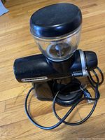 Side angle of black KitchenAid Pro Line Burr Coffee Grinder showing clear bean hopper, black lid, and motor housing on wood floor.