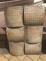 Front view of metal artwork showing six textured square panels with geometric woven patterns and varied finishes leaning against wooden stairs outdoors.