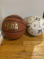 Photo showing Spalding NBA basketball and FILA soccer ball side by side on hardwood floor near white wall.