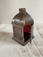 Front side view of decorative metal lantern showing detailed cut-out patterns, red glass panels, and front door opened.