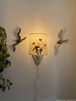 Wall sconce with the pressed leaf parchment lamp shade lit on wall with two bird wall decorations visible