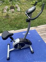 Side view of stationary exercise bike showing seat, curved handlebar, and digital monitor on blue padded mat outside.