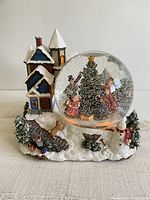 Front view of large snow globe with winter village house, snowman, dog, and family decorating tree inside glass dome filled with snow.