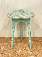 Front view of three-legged turquoise side table with clover-shaped top showing worn paint and wood exposure.