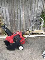 Side angle view of red Toro snowblower with black chute and white wheels against a wooden fence.