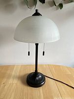 Full view of the table lamp showing black metal base, pull chains, and white glass dome shade on a wooden table with a white wall background.