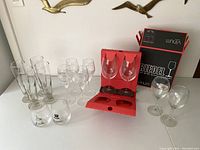 Photo showing various barware glasses arranged on a white surface against a white wall with brass bird decorations. The glasses include tall drinking glasses, small stemmed wine glasses, Foxhorn short tumblers, and two wine glasses in Riedel Vinum packaging.