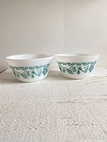 Two white milk glass mixing bowls side by side showing teal Scandinavian pattern on sides.