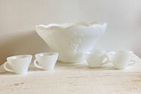 Milk glass punch bowl and four matching cups displayed on a white surface, showing the grape pattern and scalloped edge of the bowl.