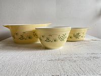 Front view of the three vintage Pyrex Shenandoah yellow dishes with green floral patterns placed side by side on a white textured surface.