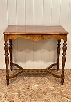 Front view of vintage wooden accent table showing scalloped apron, central carved medallion, turned legs, curved stretcher base, and wood veneer top with medium brown finish.