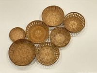 Seven woven wicker baskets of varying sizes and weave styles placed on white surface arranged in loose cluster with some overlap, showing their full round shapes and rims.