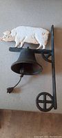 Front view of cast iron bell with white pig figure, showing overall condition and mounting bracket.
