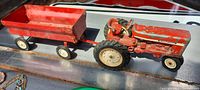 Side view of red metal vintage tractor attached to red metal wagon; tractor shows paint chipping and rubber tires; wagon rectangular bed shows wear inside.