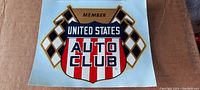 Photo of a vintage United States Auto Club decal featuring crossed checkered flags and a shield design with red and white stripes. The decal measures approximately 6 inches by 6 inches and is unused.