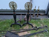 Larger pair of black cast iron andirons with brass finials standing next to wrought iron fireplace tools including shovel, poker, tongs, and smaller wrought iron andirons.