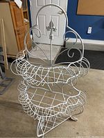 Front perspective of the white wrought iron French plant stand showing two wire basket shelves and arched back with fleur-de-lis detail.