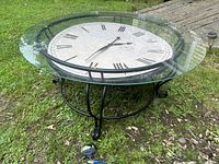 Front angle showing the clock face beneath the clear glass table top and the black metal base on grass.