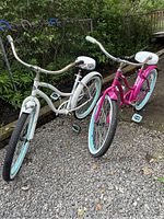 Two Huffy cruiser bikes side by side on gravel; white adult and pink teen/small adult size bikes visible with turquoise rims and pedals, white seats, and upright cruiser handlebars