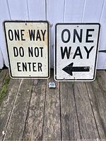 Both street signs displayed upright on wooden surface against a white wall, showing overall size and condition including dirt, scuffs, and mounting holes.