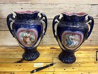 Pair of cobalt blue porcelain vases with ornate handles and painted Victorian scenes placed on wooden surface with pen for scale