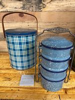 Vintage blue plaid metal insulated cooler beside stacked blue speckled enamelware bowls in metal rack, placed on wooden surface with wooden backdrop.