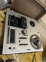 Top and perspective view showing Akai branded tape head cover, analog VU meters, control knobs and tape reels on metal casing with wooden sides.