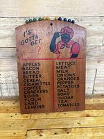 Front view of wooden grocery reminder board with item list and painted figure, showing colored pegs at top.