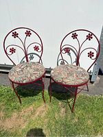 Pair of red wrought iron chairs with chippy paint and floral metal backs, each with a round patterned fabric cushion, shown outdoors against a white wall.