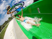 Person sliding down a bright green water slide at a waterpark with multiple taller slides in background under blue sky.