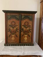 Front view of wooden medicine cabinet with hand-painted floral artwork on two doors, green trim and reddish accent lines.
