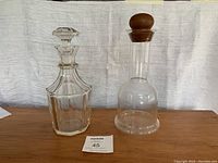 Two decanters side by side, one crystal with faceted stopper and one smooth glass with wooden stopper, on wooden surface with fabric backdrop.
