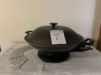 Black electric wok with domed lid and knob, sitting on table next to chrome grill tray and power cord