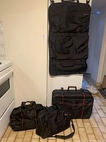 Main set of four black cloth bags: large wheeled luggage, garment bag hanging on wall, two smaller carrier bags on floor