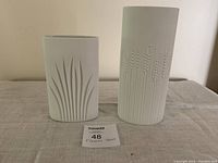 Two white Rosenthal vases on light fabric. One is taller cylindrical vase with dotted and lined texture. Other is shorter, curved rectangular vase with raised leaf patterns. Small inventory paper in front.