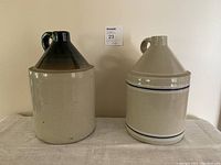 Photo of two ceramic jugs on beige cloth against plain wall, showing front view of both with dark top jug on left and striped jug on right
