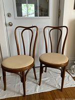 Front view of two matching wooden side chairs with rounded beige recovered seats, curved backs.