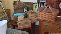 Photo showing a group of five baskets on and around a small table in a room including two picnic baskets with wooden handles and three woven storage baskets of various sizes and shapes.
