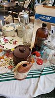 Photo showing vintage glass bottles, ceramic crocks, floral bowls, brown ceramic crock, and glass jars on a table with a floral tablecloth.