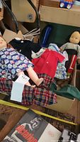 Box containing multiple vintage dolls dressed in various outfits including floral dresses and coats, displayed for size and content.