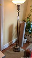 Metal floor lamp with frosted glass square shade, metal stem, and marble base having wear and damage. Lamp stands next to rolled carpet and plant against wall.