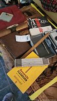 Photo showing a collection of vintage books, the Polaroid Brownie Flash Holder in its original yellow box, wooden and metal measuring sticks, and a folded ruler.