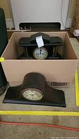 Box containing a black wooden mantel clock with columns and an arched black wooden mantel clock on the floor.