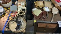 Lot shown on two surfaces with vintage hair dryers, vintage necklace on mirrored tray, ornate hand mirrors and picture frames, electric shaver and razor on wooden chest and nearby table.