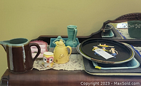 Overview photo showing multiple vintage ceramic pitchers, a chick egg coddler, and colorful decorative dishes and trays laid on a wooden surface with a lace runner.